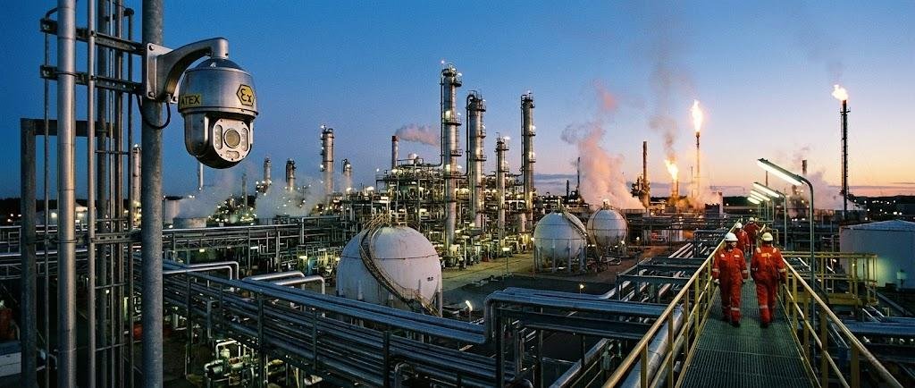 SWGJ stainless steel explosion-proof PTZ camera monitoring a petrochemical plant with spherical storage tanks and distillation towers at dusk.